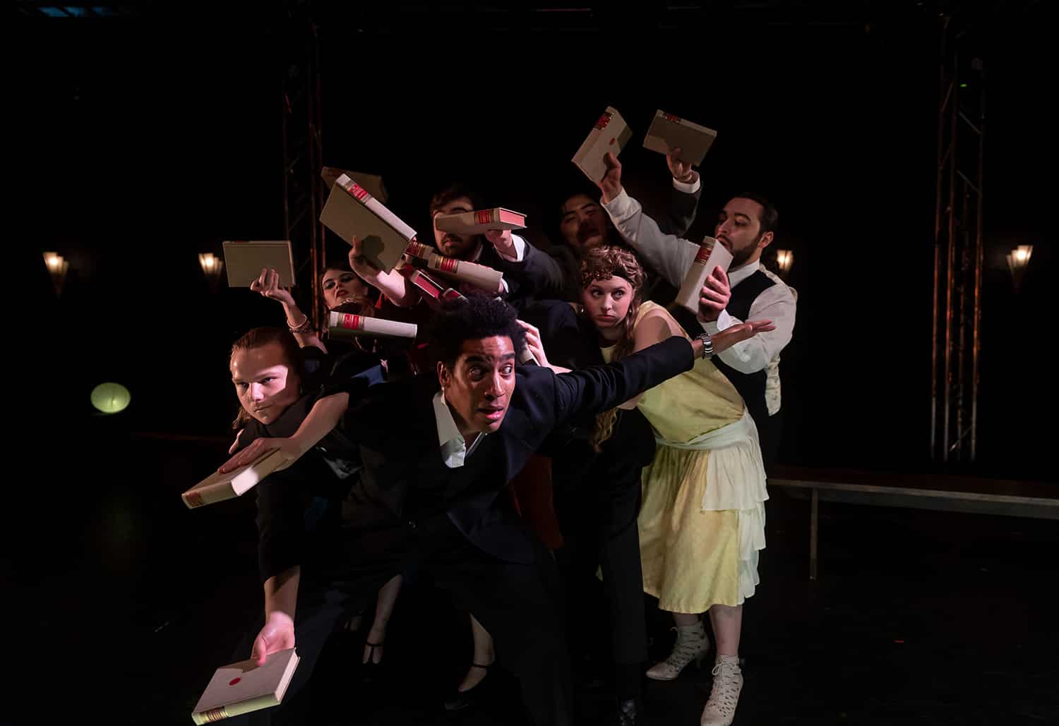 A group of actors clustered on stage carrying a book each