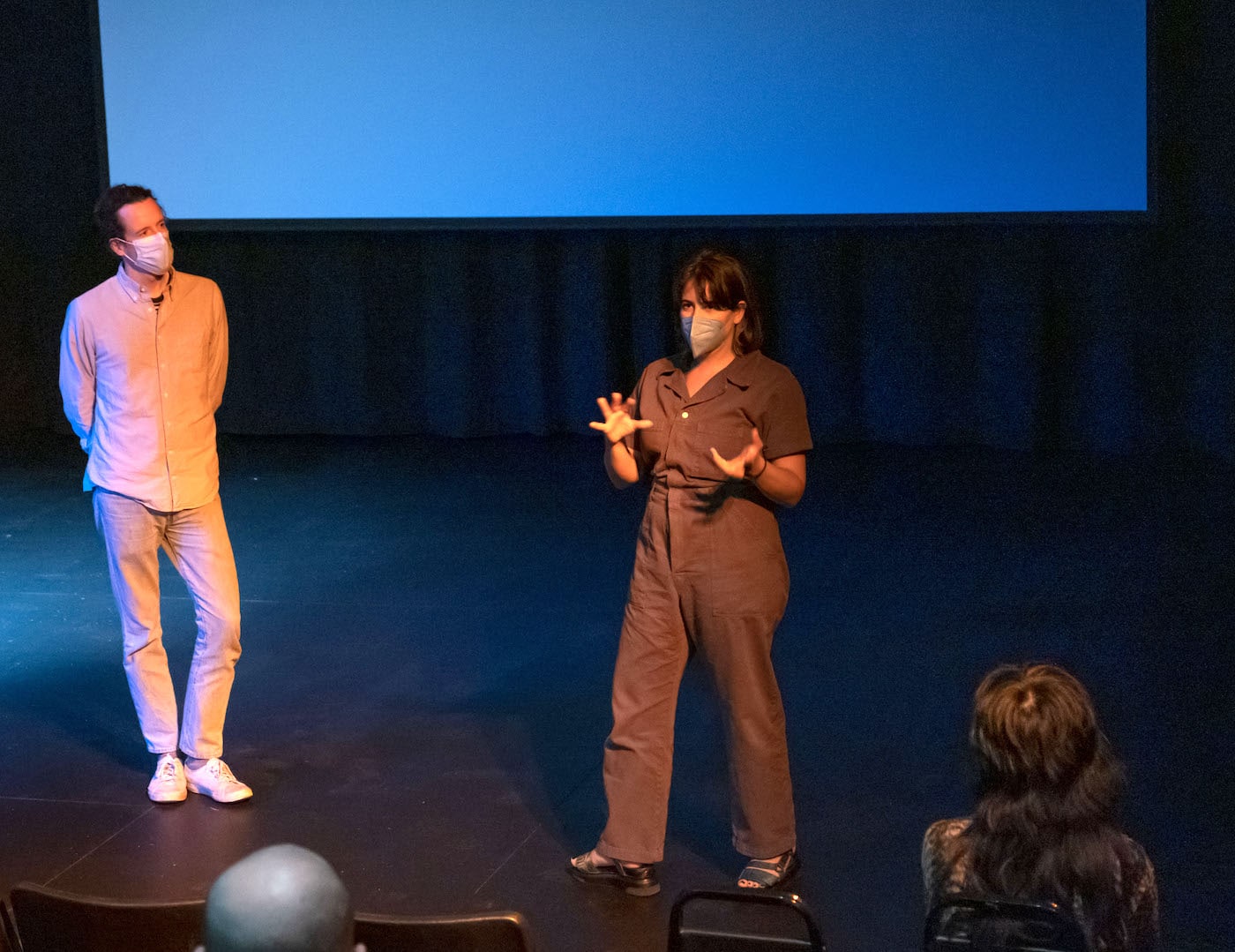 Two people in masks talk from a stage.