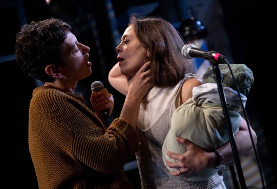 Actor holds microphone in one hand while the other holds the neck of another actor with a baby bundle