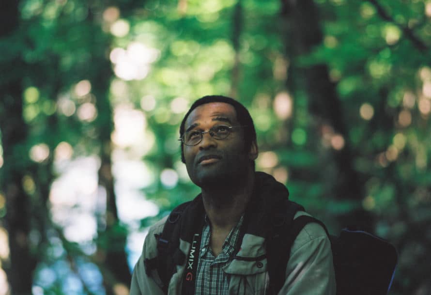 Man wearing backpack and camera slung over neck looks up at forest