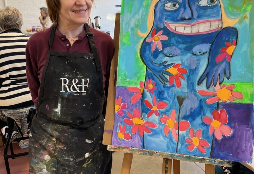 Cynthia Pepper stands in painter smock next to easel with painting of blue figure surrounded by flowers