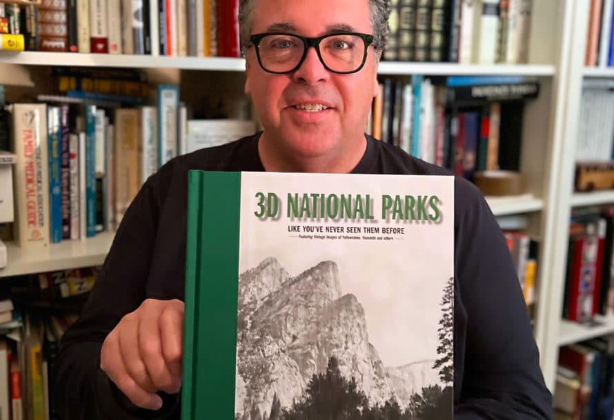 David Bossert stands against bookshelf holding book '30 National Parks Like You've Never Seen Them Before'