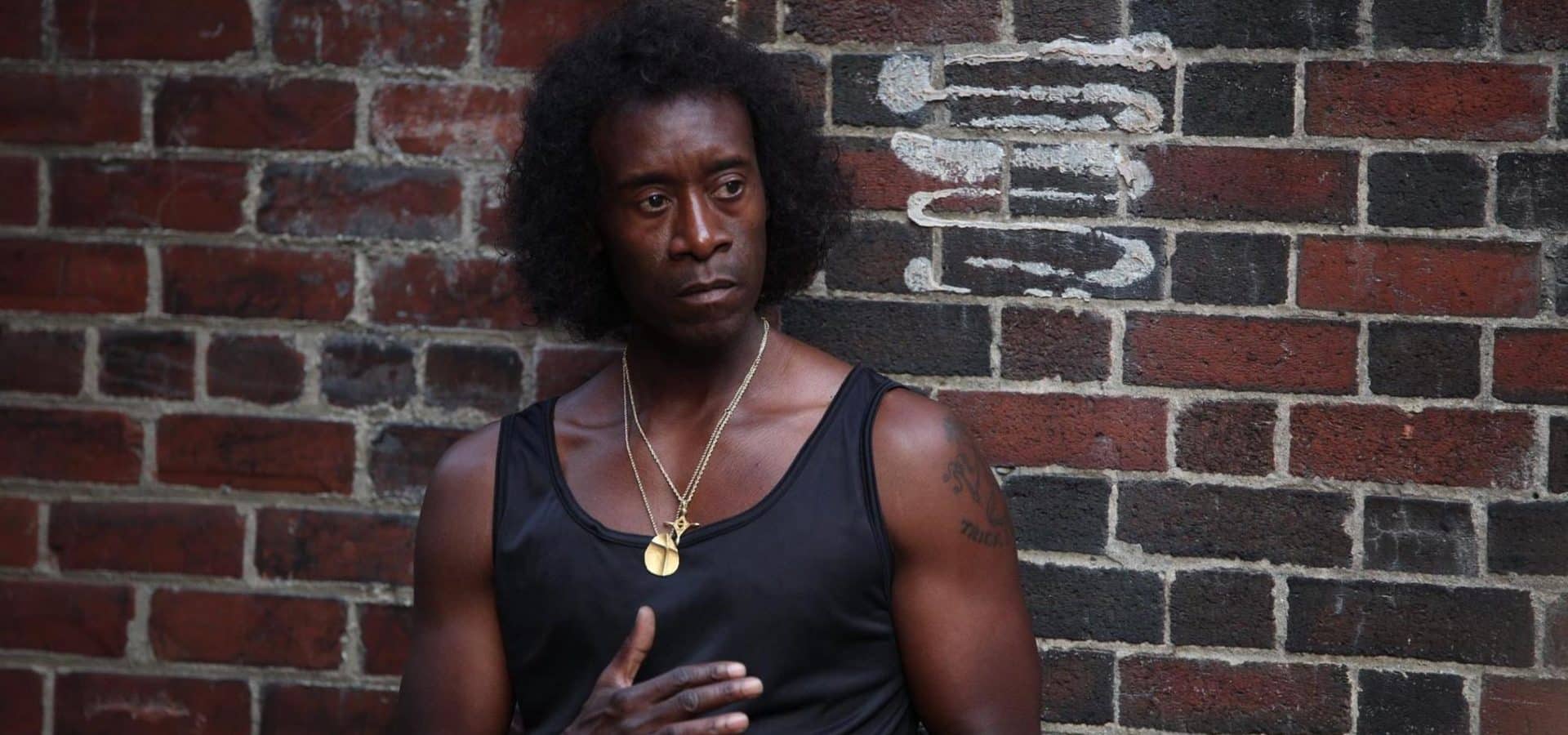 Don Cheadle as Miles Davis, in a black tank top, standing against a brick wall.