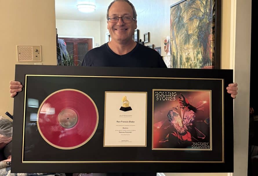 A man in glasses holds up a framed award from the Grammys for performing on a Rolling Stones recording.