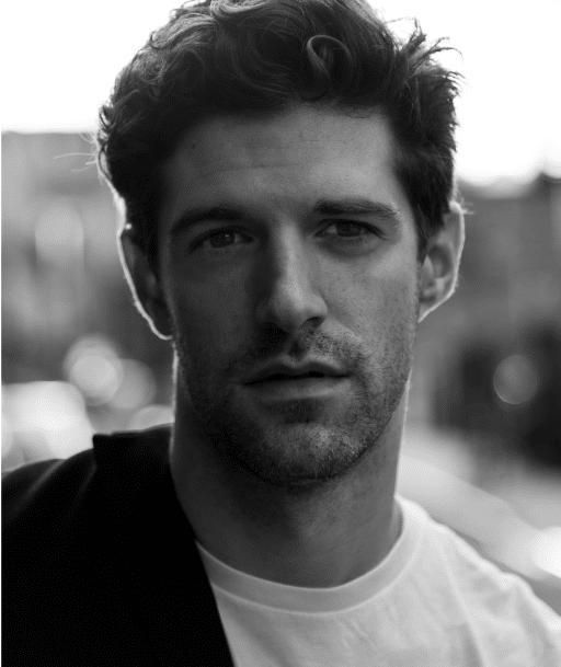 Black and white headshot of man in white T-shirt with a jacket hanging on his right shoulder