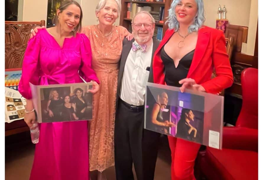 A man in a tux stands next to three women in dresses. Two of them are holding up photos of themselves performing.