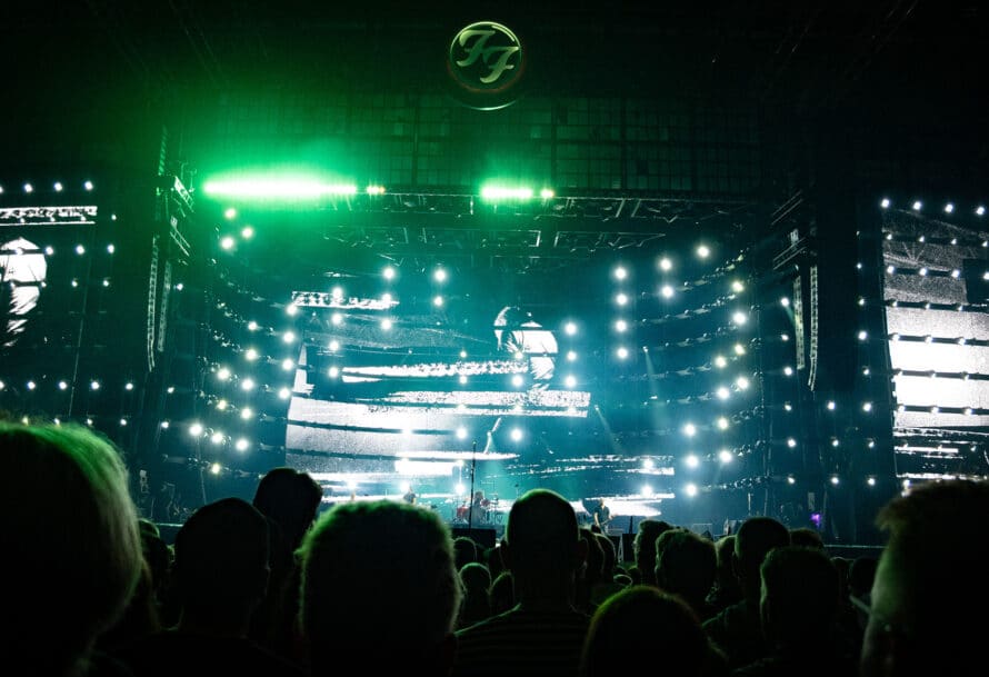A concert scene featuring an audience in front of a brightly lit stage, showcasing a large screen with black and white visuals and vibrant green lights.